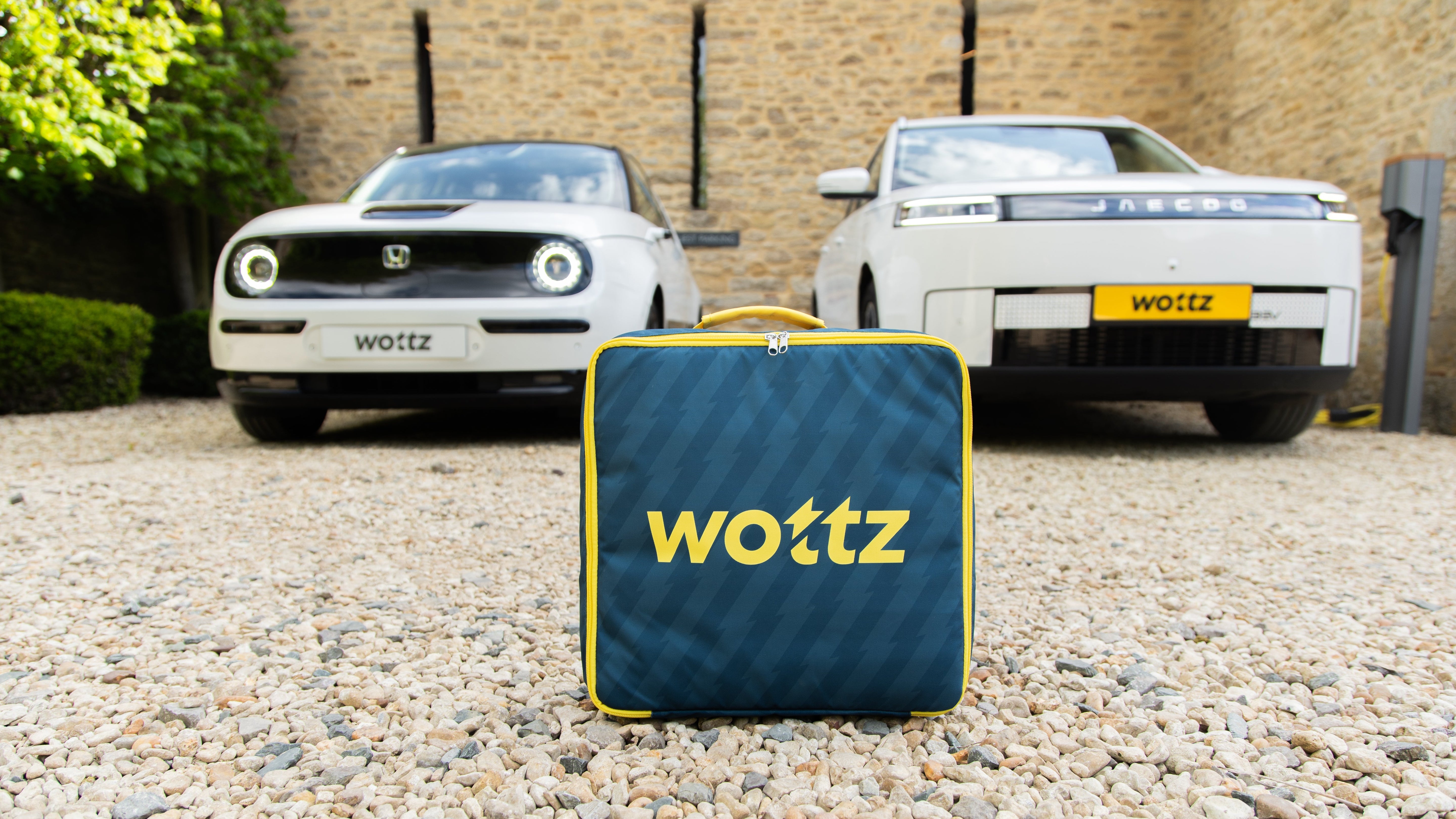 Wottz branded blue carry bag with yellow logo on gravel driveway with Honda e and Jaecoo SUV in the background