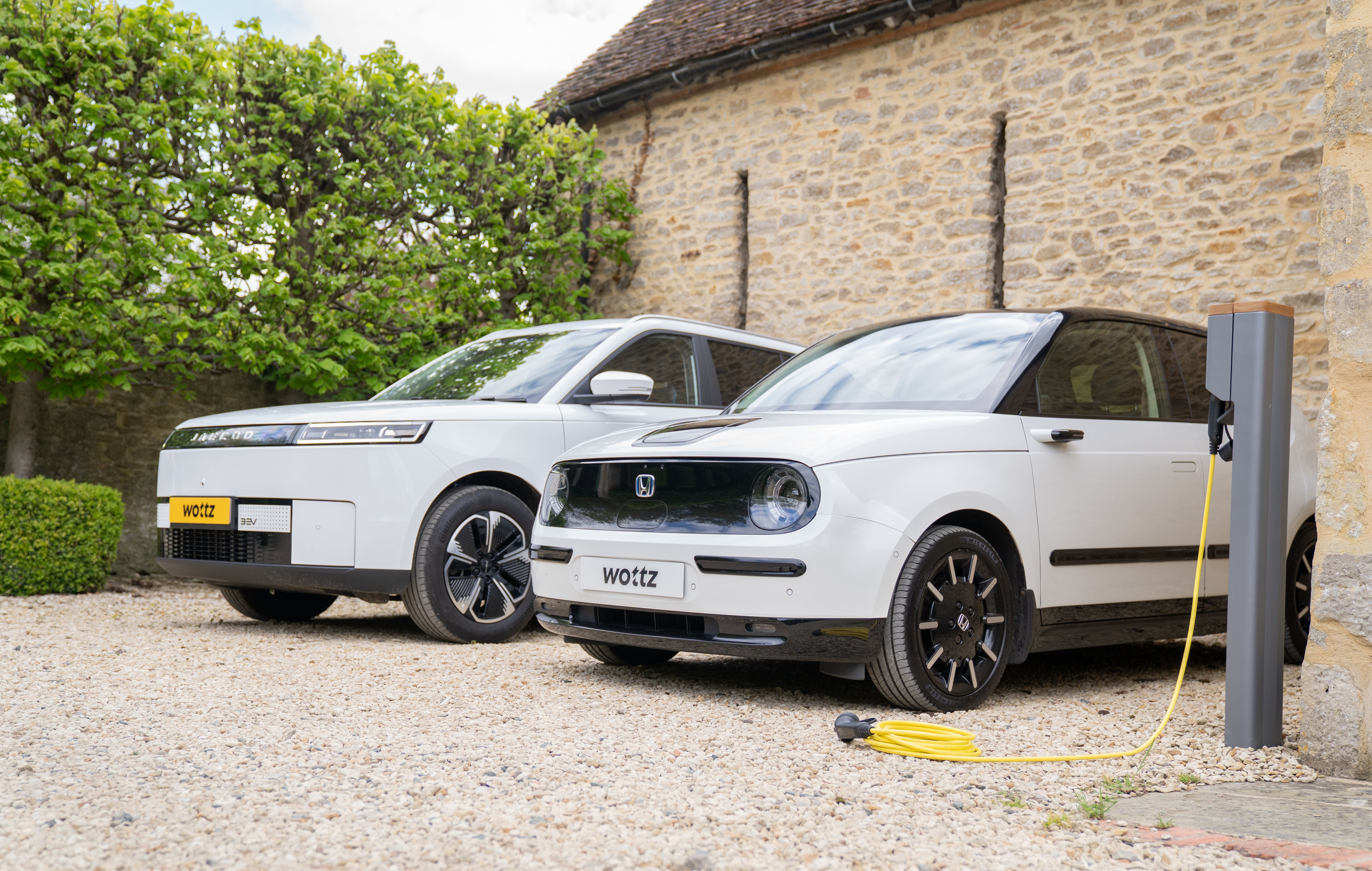 Two white Wottz-branded EVs parked at a Cotswolds cottage with the Honda e charging from a wall post via yellow cable
