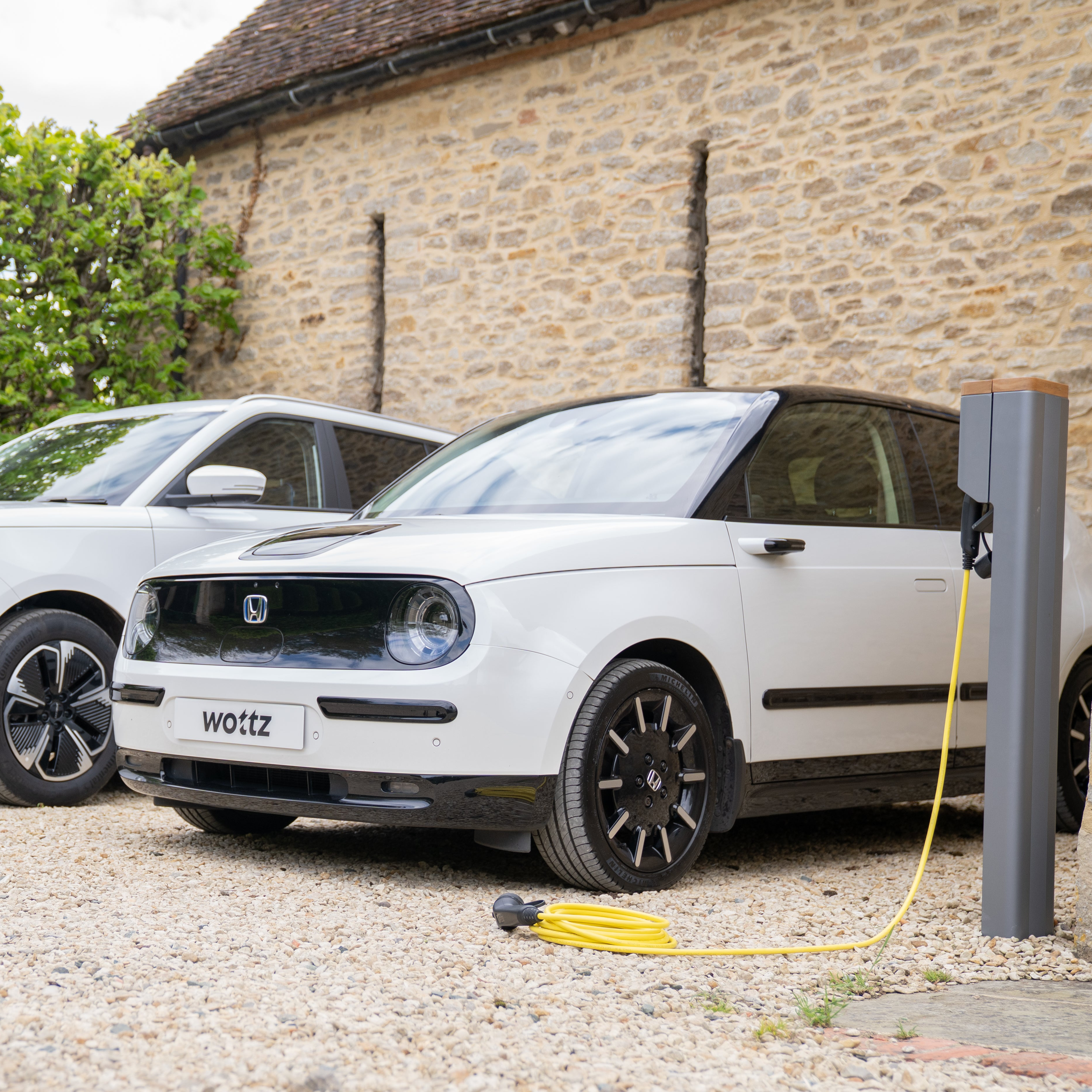 White electric car with 'Wottz' branding charging at a charging station in front of a stone building.