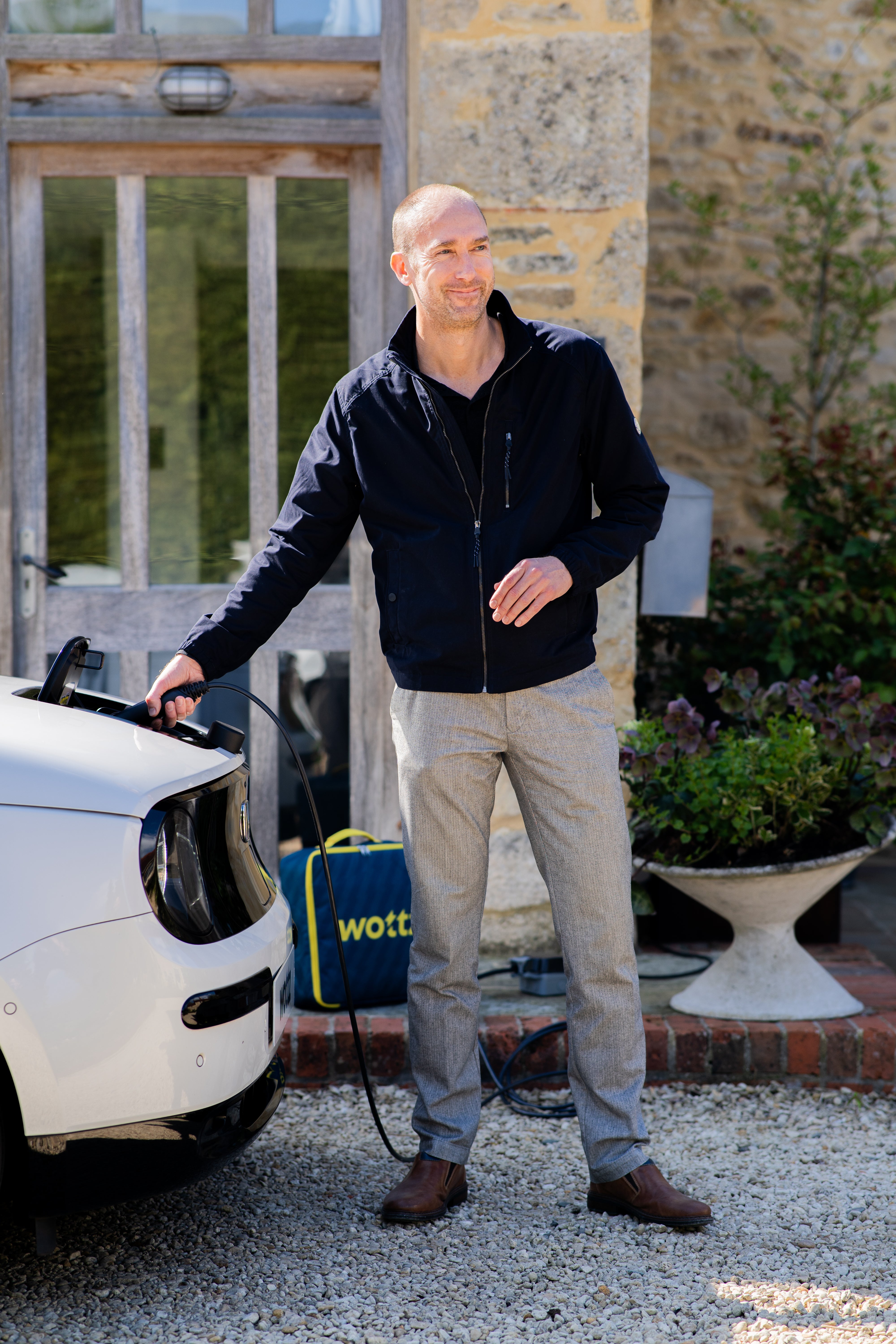 Man connecting a Wottz portable charger to a white Honda e with a carry bag on the ground at a Cotswolds stone house