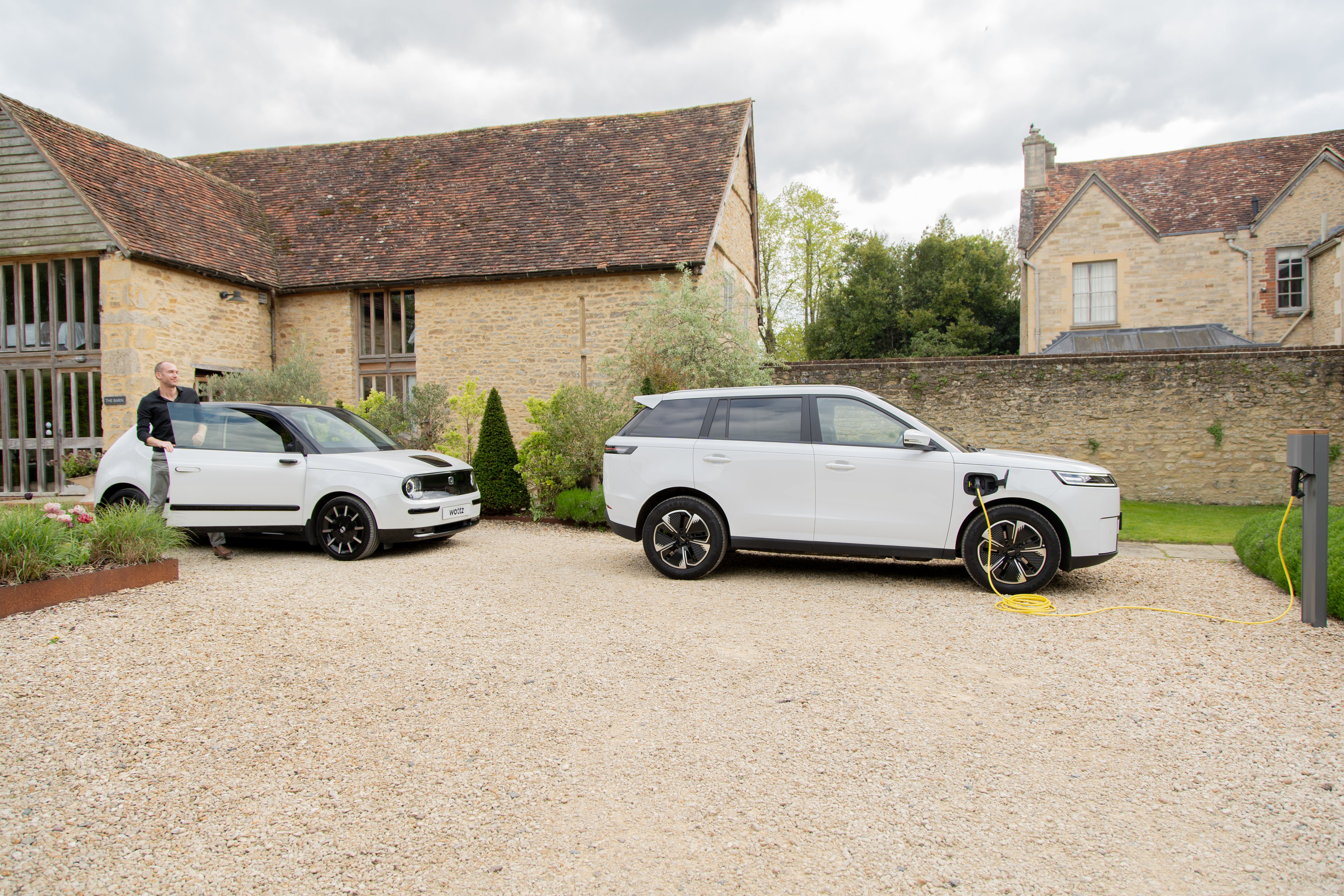 White Honda e and Jaecoo SUV at a Cotswolds barn with the SUV charging from a wall post via yellow Wottz cable