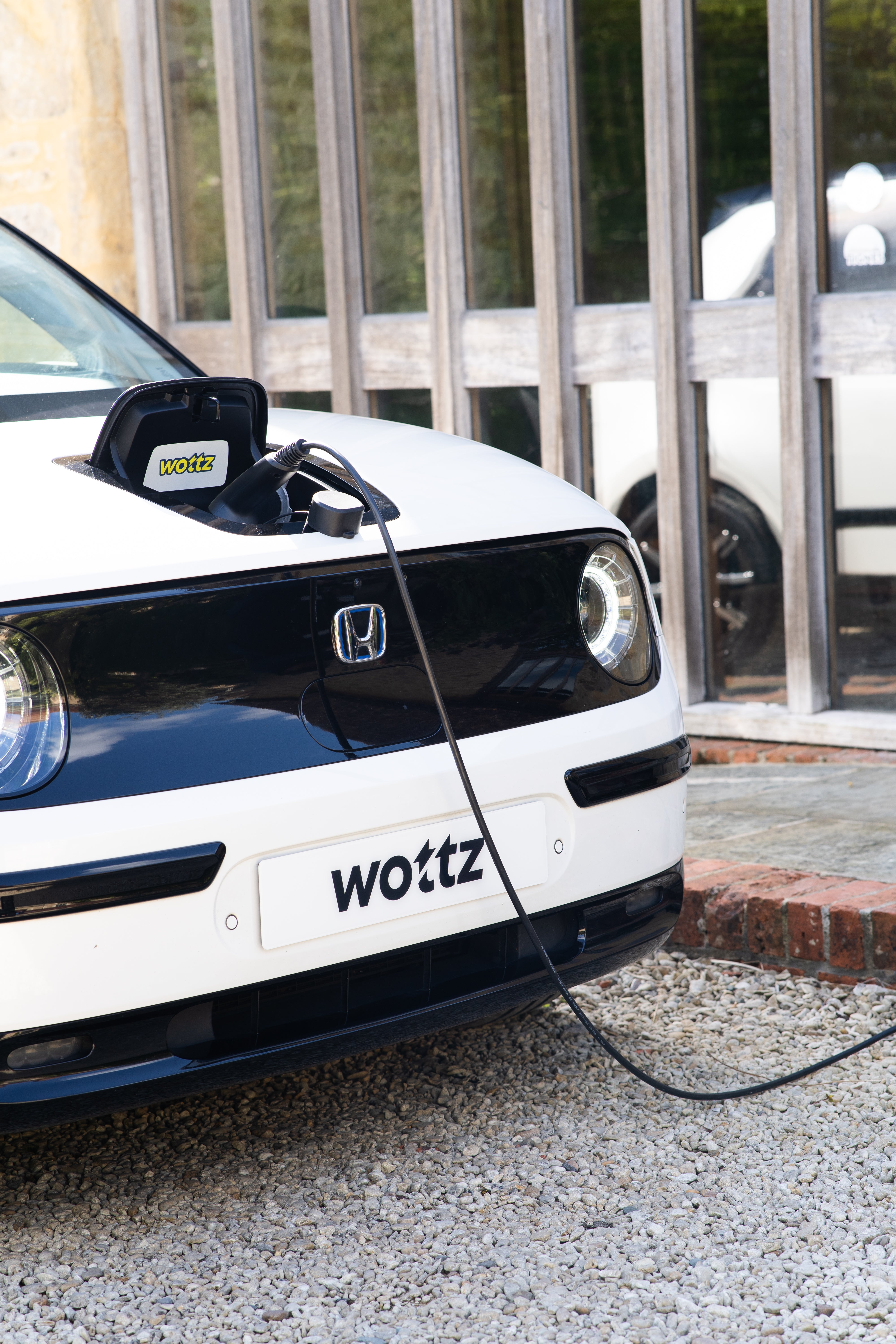 White Honda e charging at a Cotswolds stone barn using a Wottz portable charger with carry bag visible