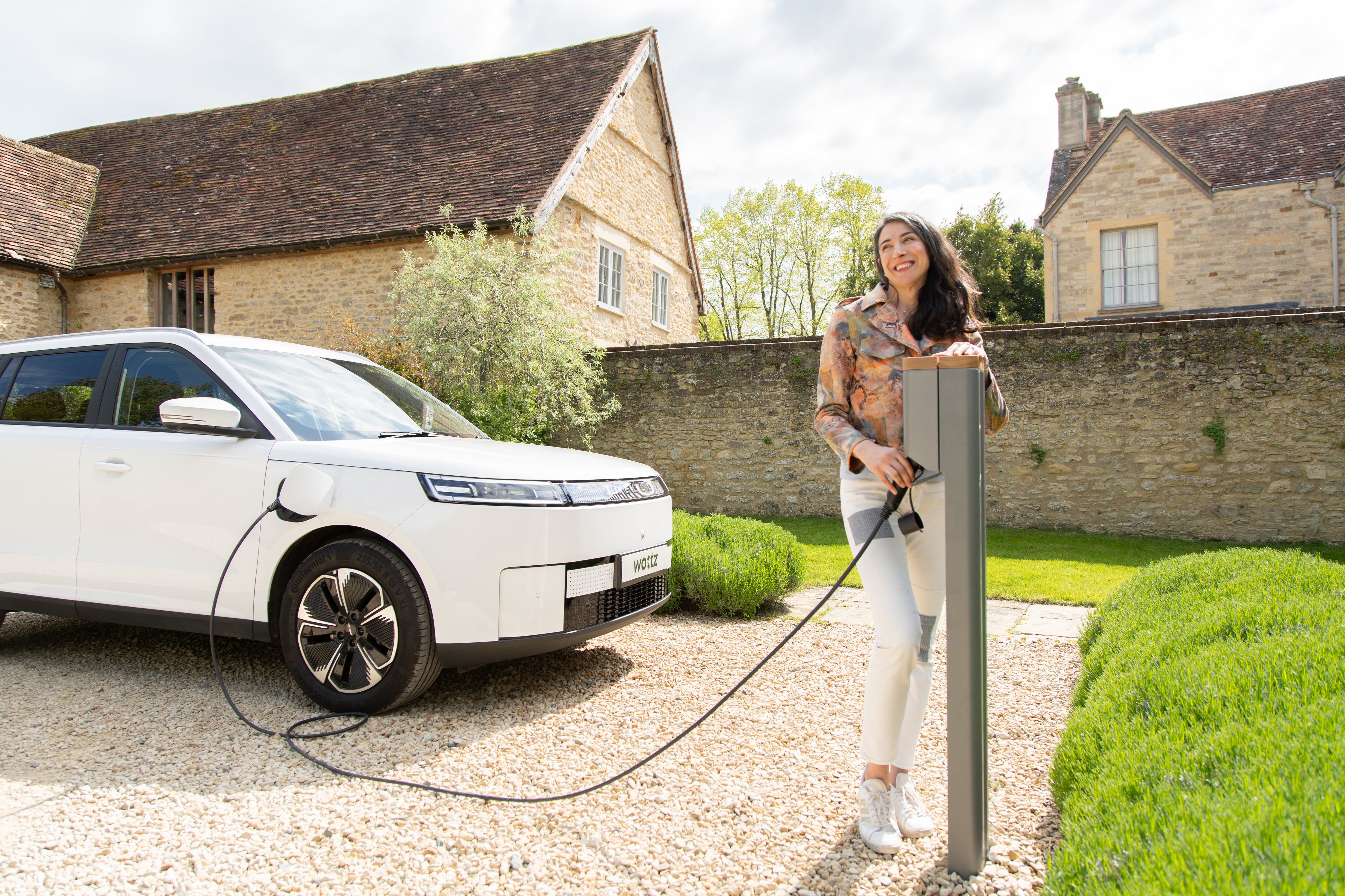 Woman connecting a Wottz charging cable at a freestanding charge post beside a white electric SUV