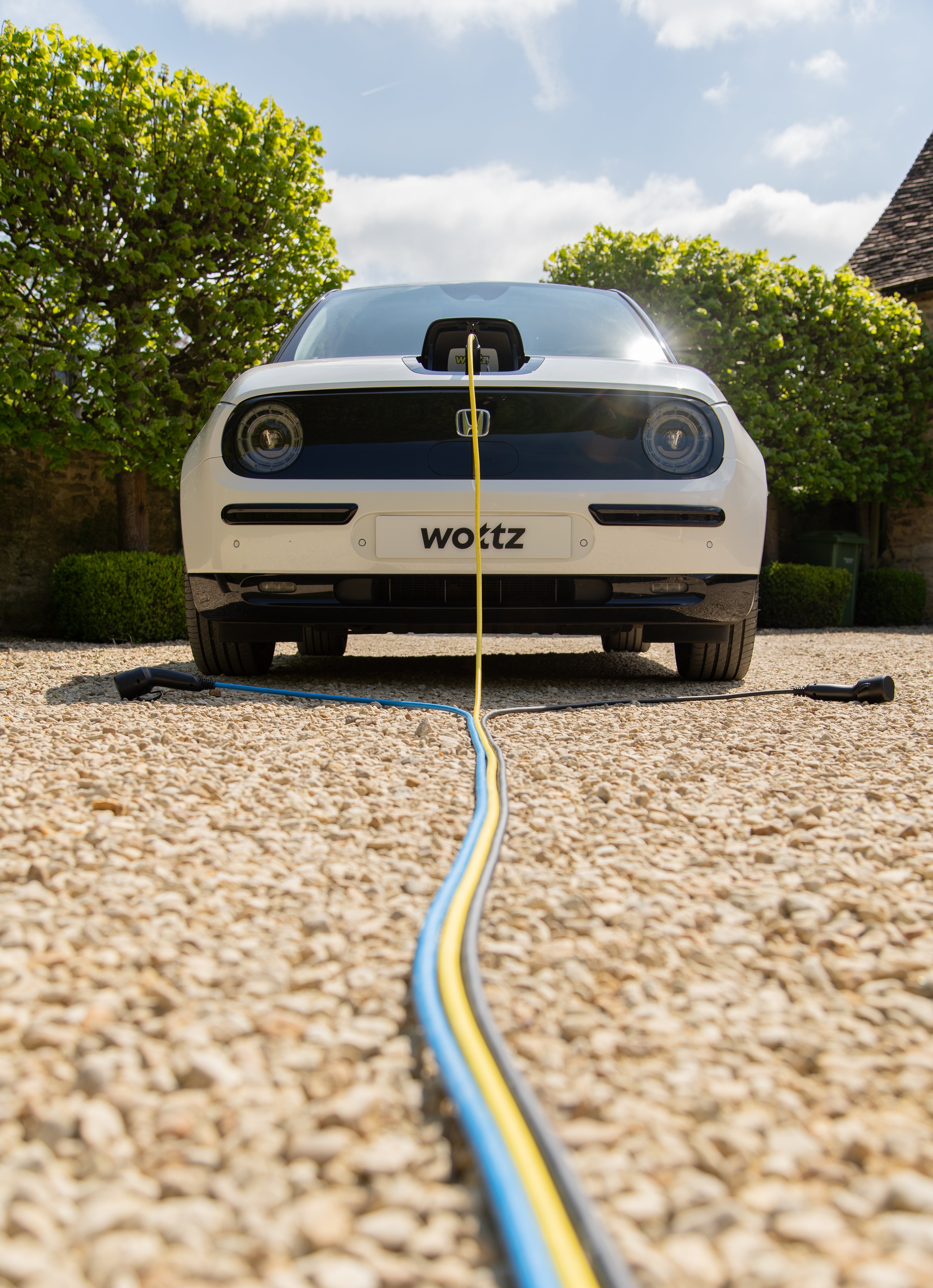 Front view of a white Honda e charging with blue and yellow Wottz cables on a gravel driveway