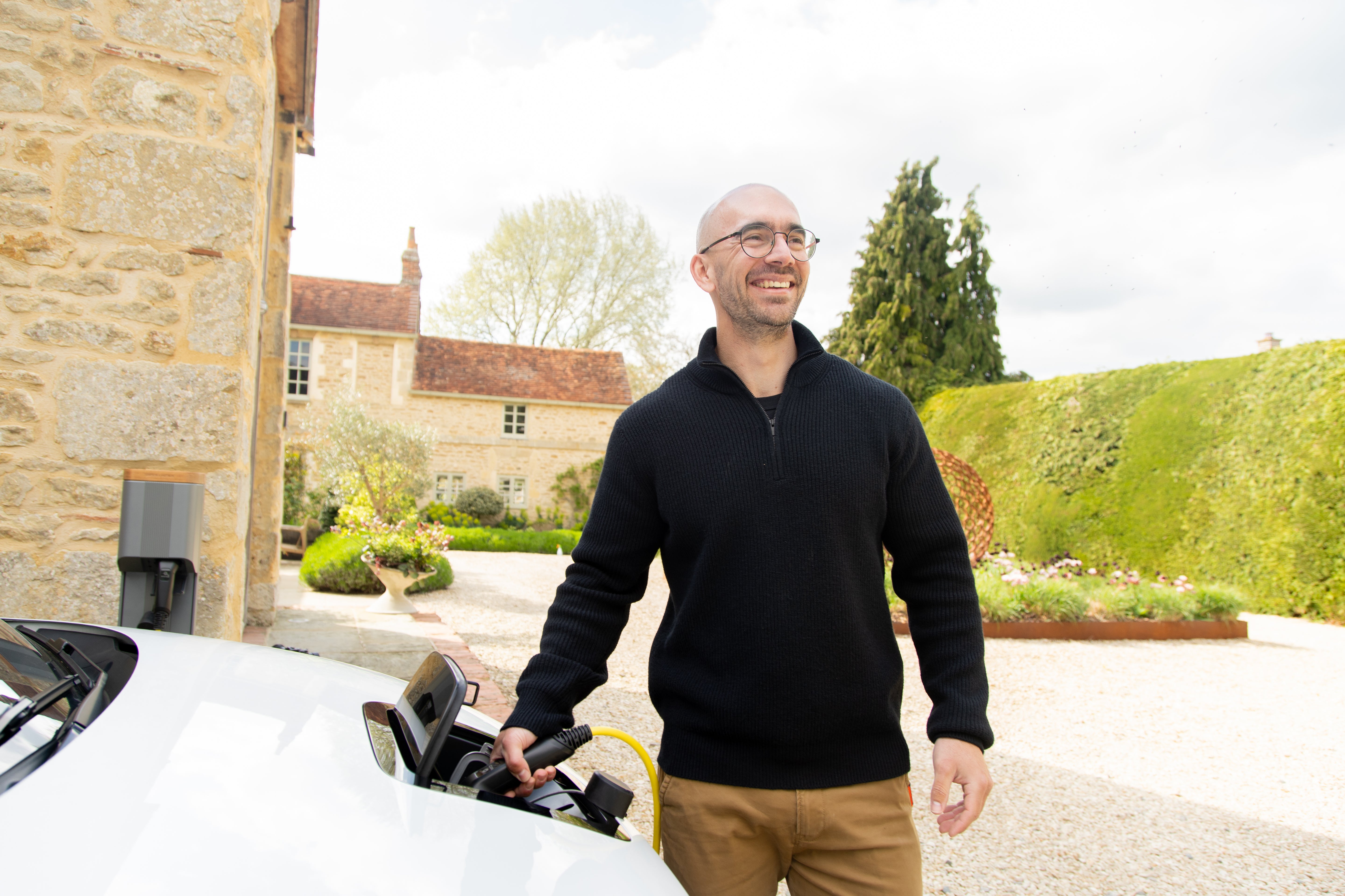 Man plugging a yellow Wottz EV charging cable into a white electric car at a Cotswolds stone house