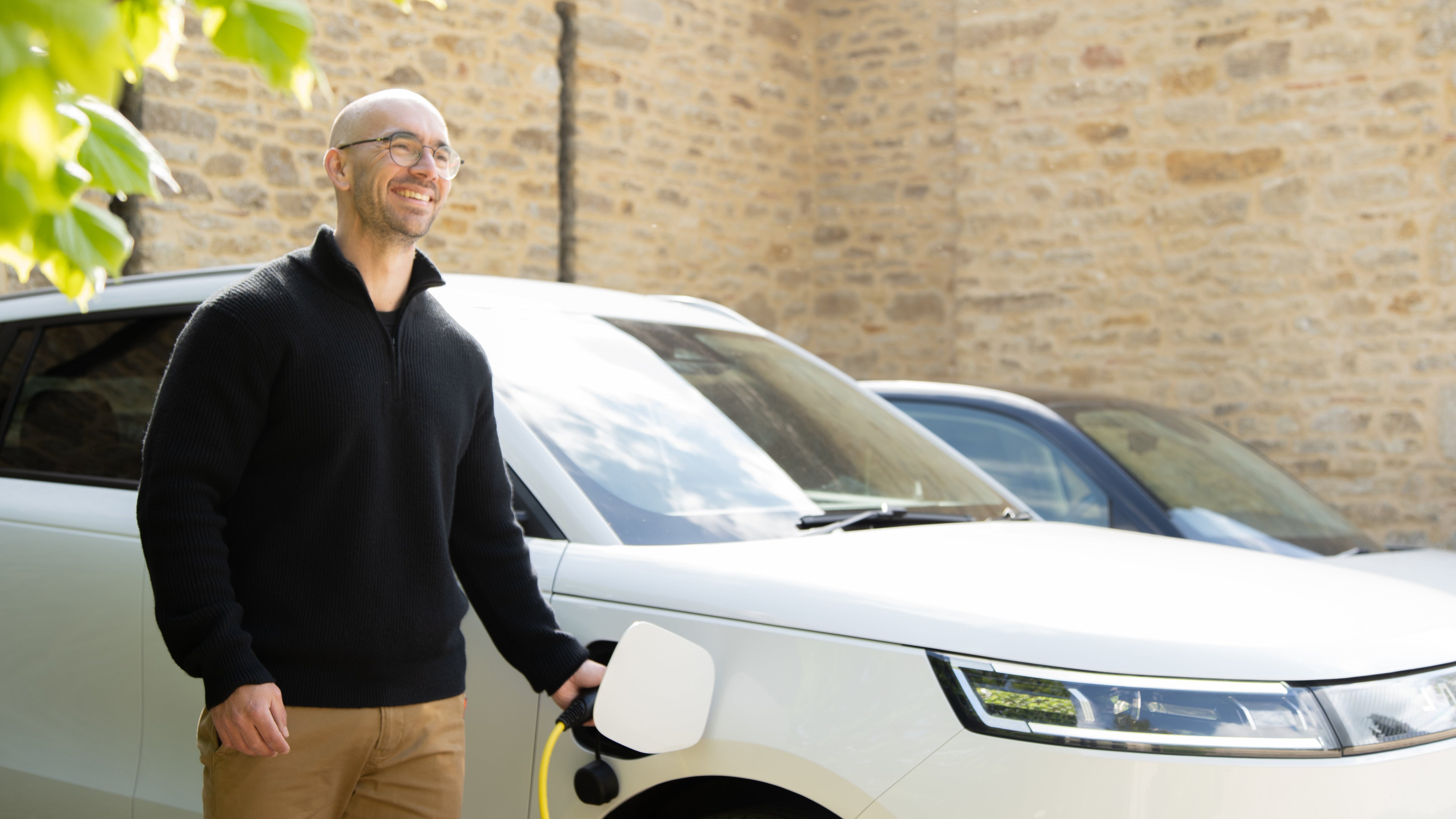 Man with glasses smiling while plugging a yellow Wottz cable into a white Range Rover at a Cotswolds stone wall