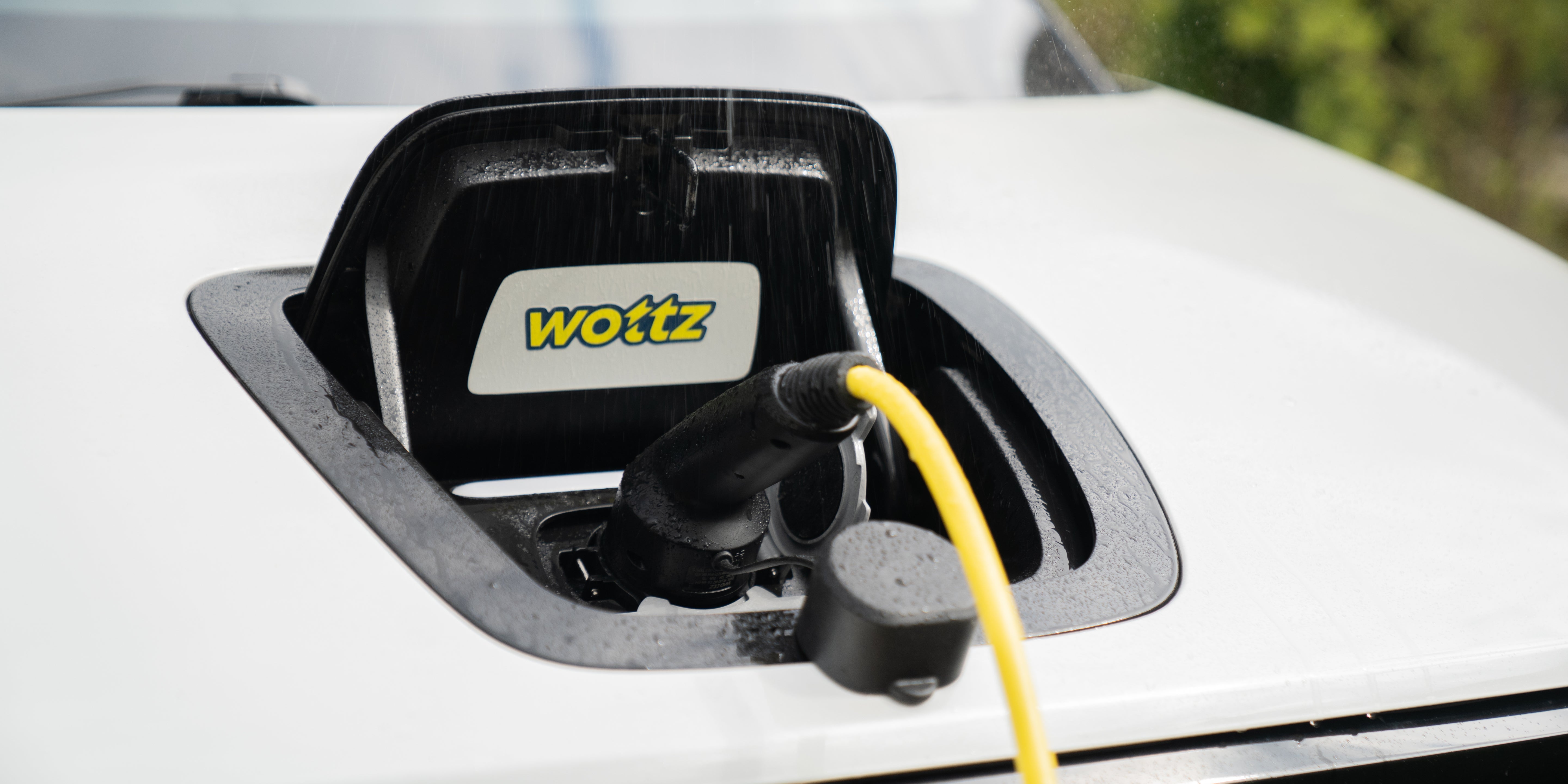 Close-up of a yellow Wottz cable plugged into a white car charge port with Wottz logo badge and rain drops